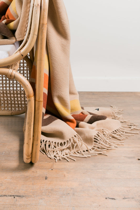 Beige blanket with fringe draped over a wicker chair on a wooden floor.