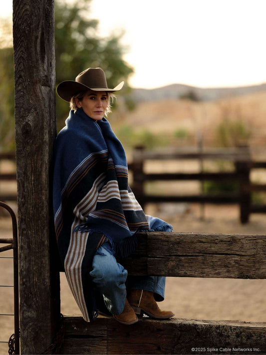 Person wearing a cowboy hat and blanket sitting on a wooden bench outdoors.