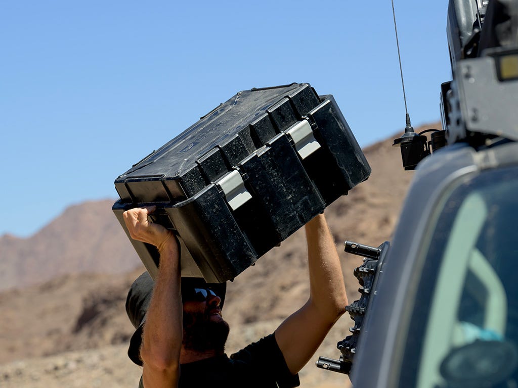 Person carrying a large black toolbox over their head in a desert setting with a vehicle in the background.