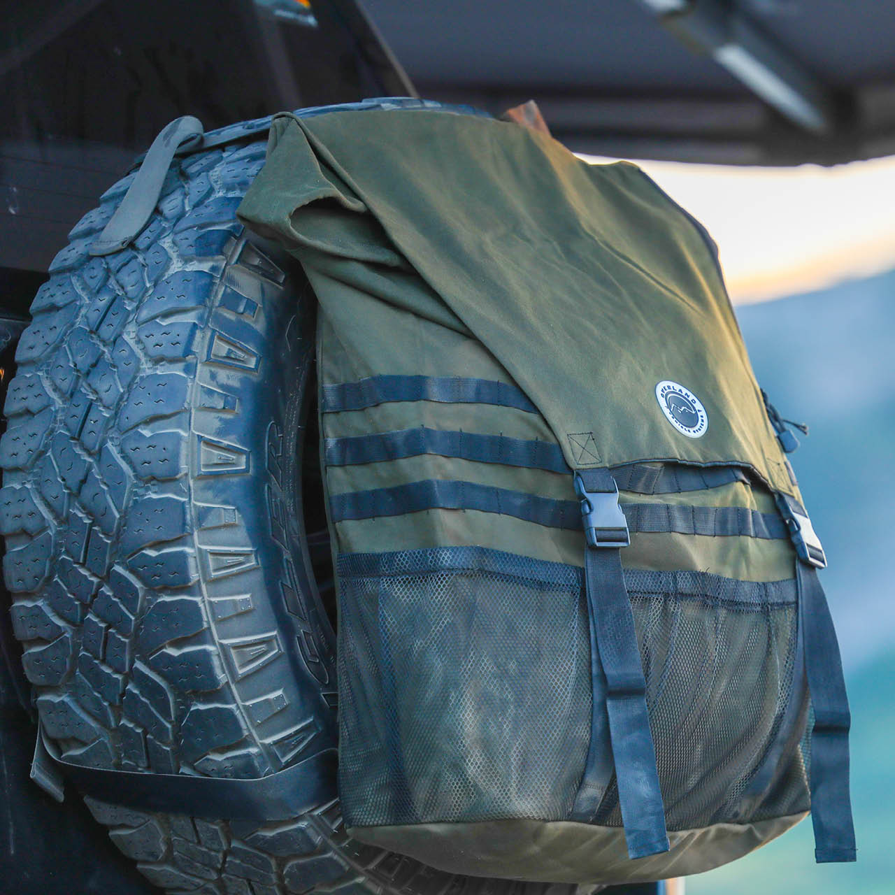 Green canvas trash bag with a logo attached to a tire, with a blurred natural background