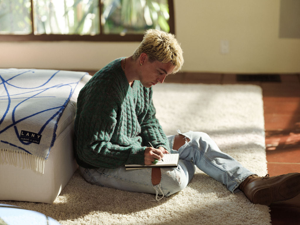 Person sitting on a carpeted floor writing in a notebook, wearing a green sweater and light blue jeans.