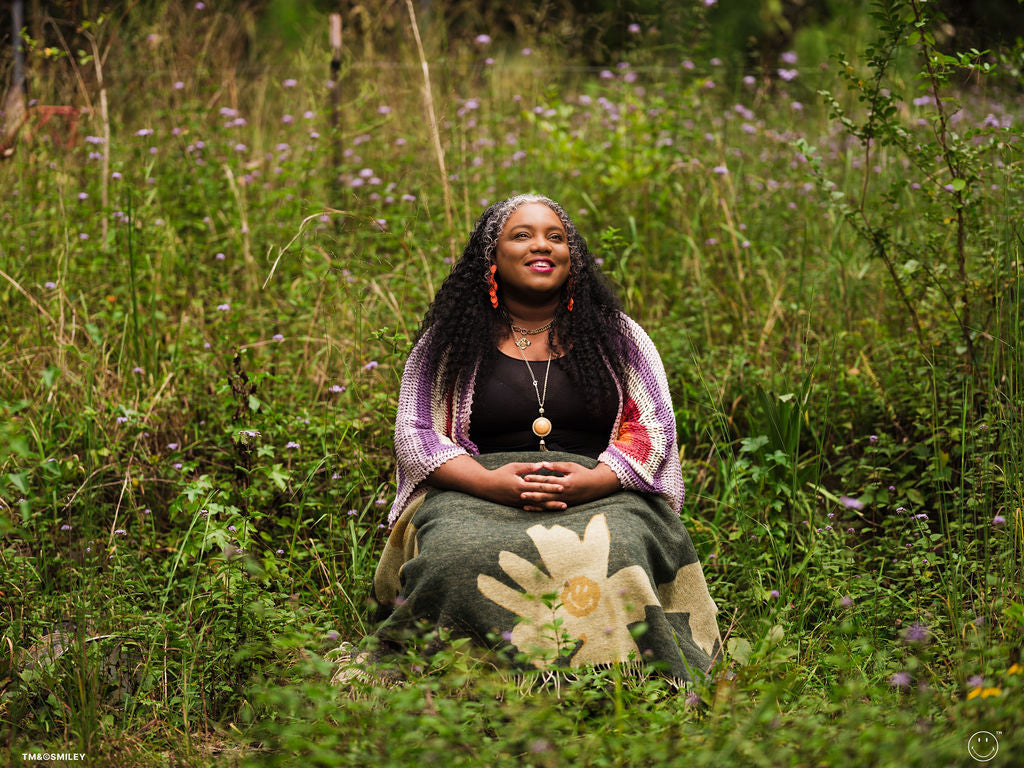 Woman sitting in a field of tall grass and wildflowers, wearing a colorful shawl and green skirt.
