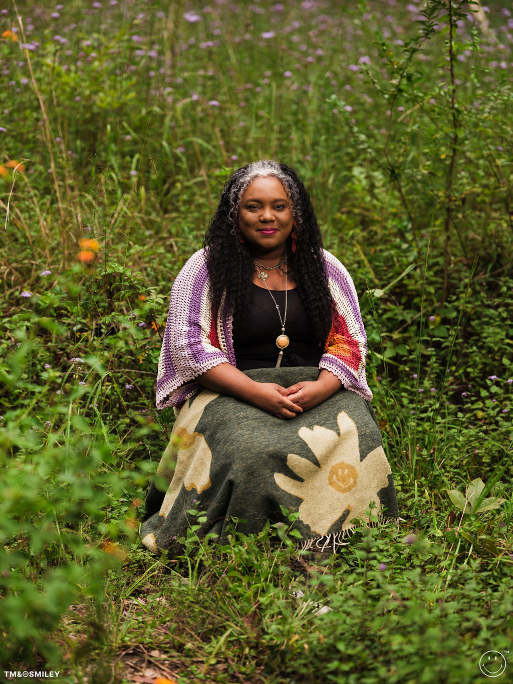 Woman sitting in a field of flowers wearing a colorful shawl and patterned skirt.