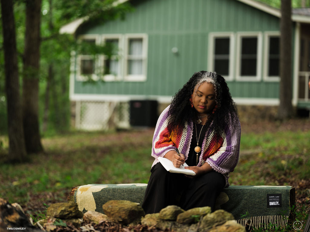 Woman sitting outdoors with a notebook, wearing a colorful sweater, with a green house in the background.