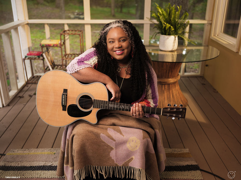Woman holding a guitar on a wooden deck with a plant and table in the background