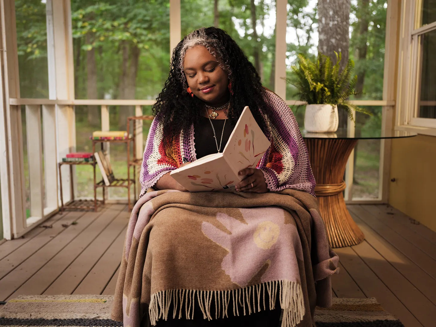 Woman reading a book on a wooden deck with a blanket and decorative items.