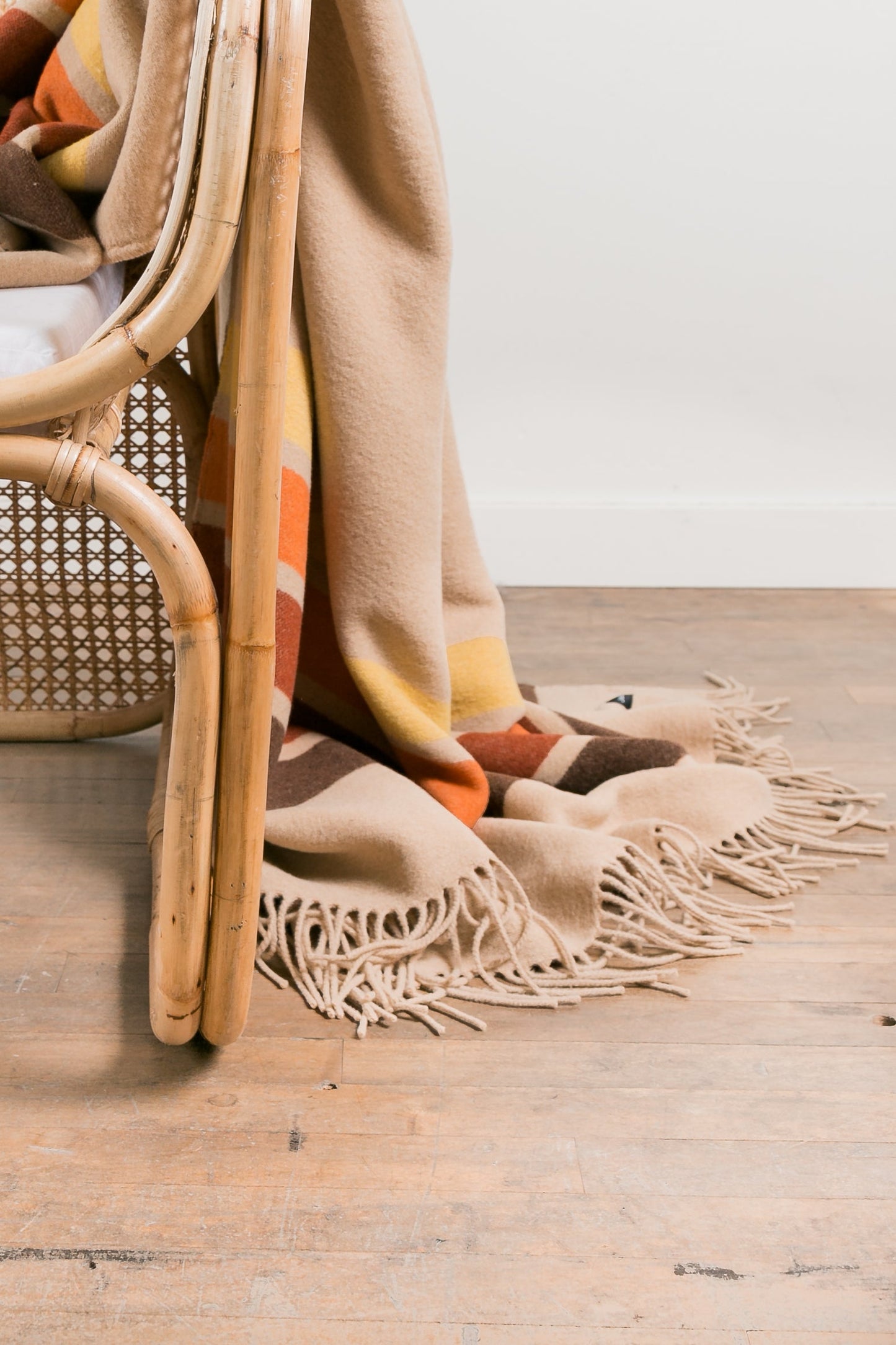 Beige blanket with fringe draped over a wicker chair on a wooden floor.