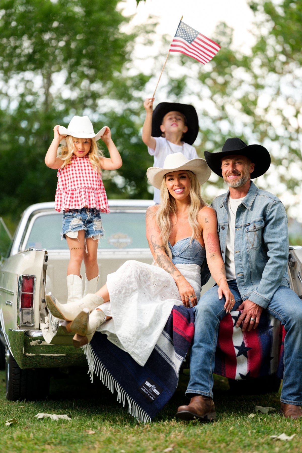 Family with children in cowboy hats sitting on a truck bed with an American flag, outdoors.