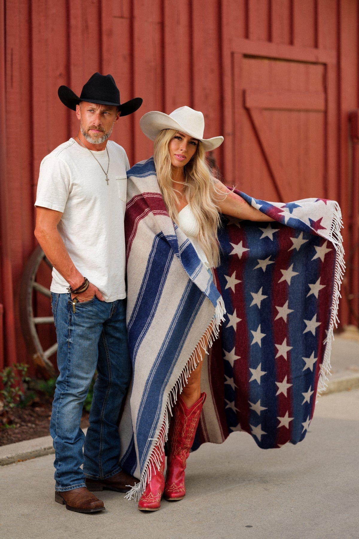 Man and woman in cowboy hats with a colorful blanket featuring stars and stripes, standing in front of a red barn.