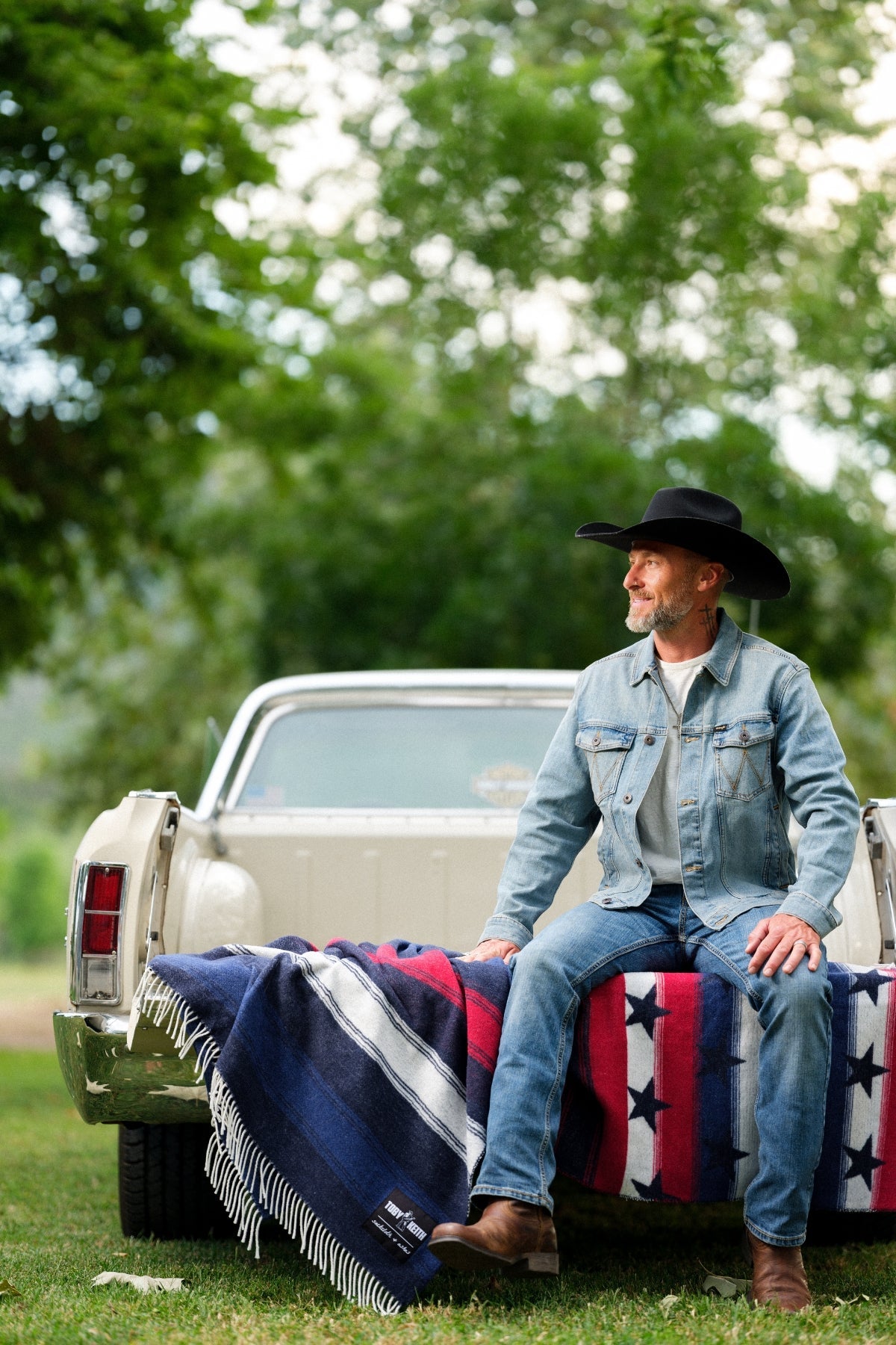 Man in a cowboy hat sitting on a truck bed with a flag blanket, surrounded by greenery.