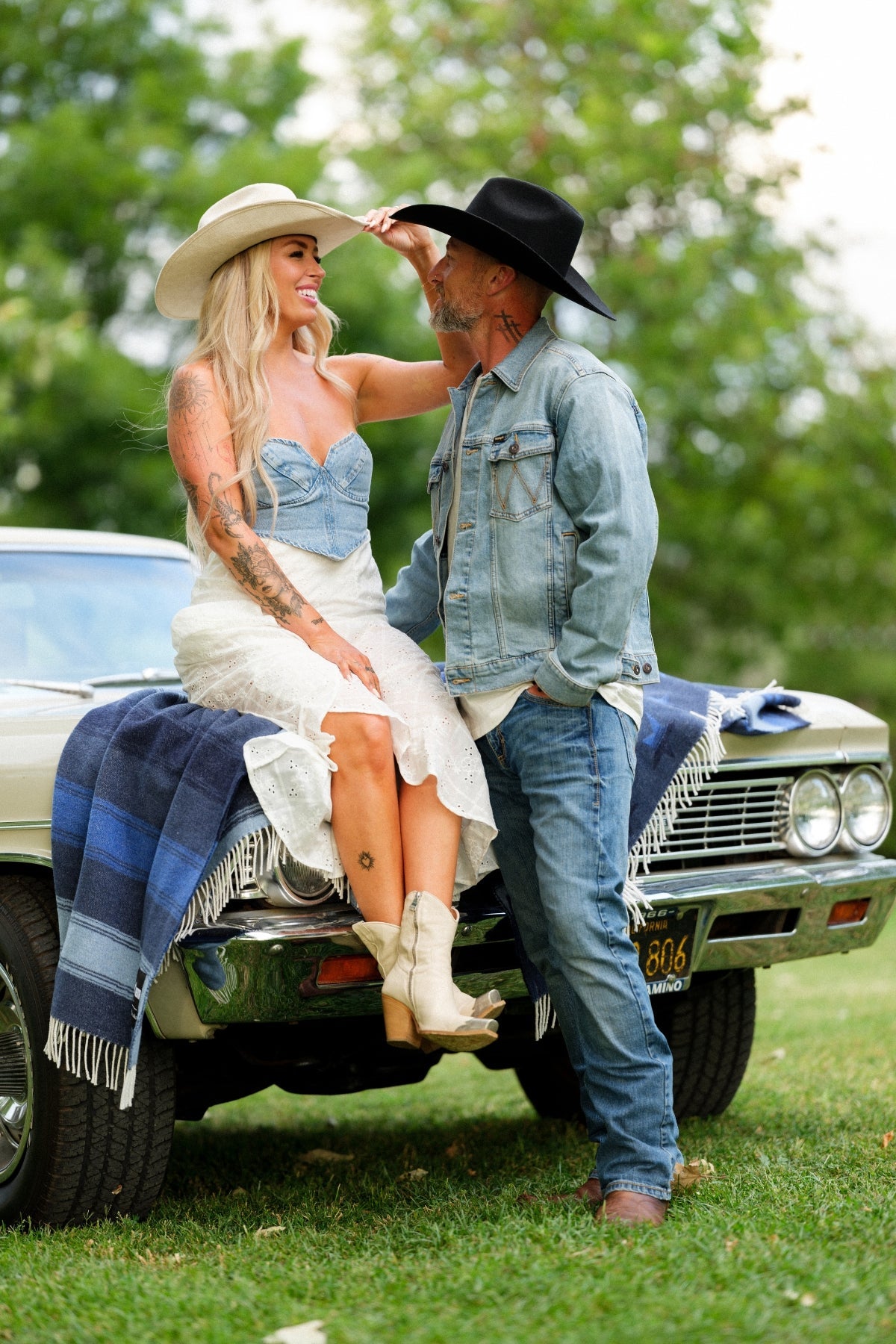Couple in cowboy hats sitting on a classic car in a grassy area with trees in the background