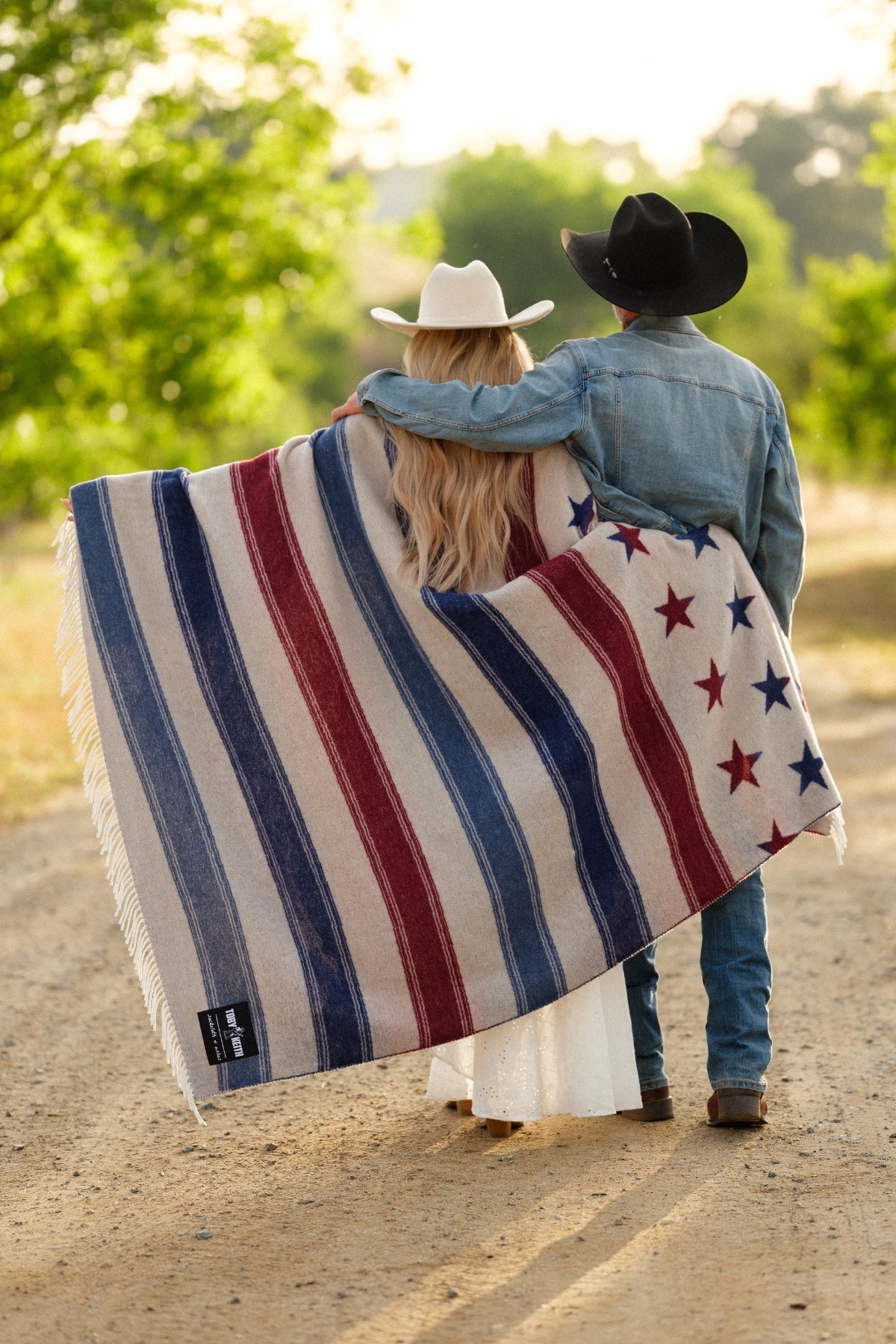 Two people wrapped in a red, white, and blue striped blanket with star patterns on a dirt road.