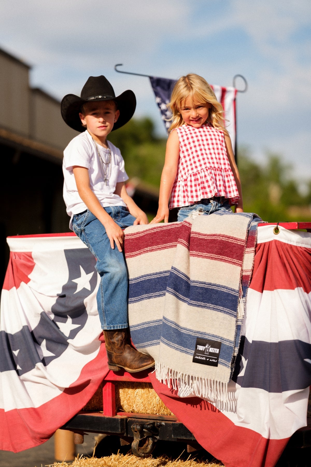 Two children sitting on a cart with a patriotic blanket, one wearing a cowboy hat.