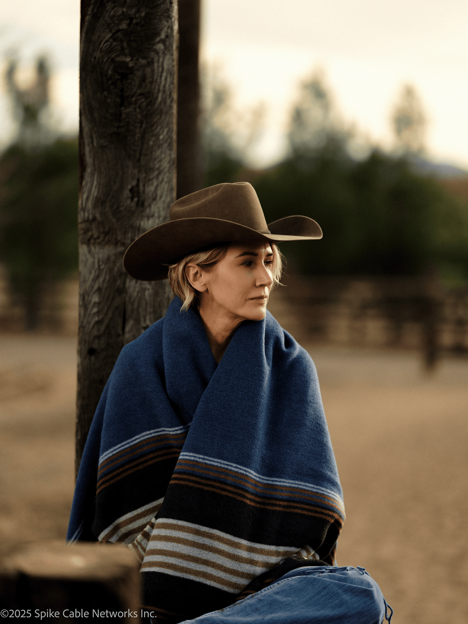 Person wearing a cowboy hat and blue poncho sitting against a tree in a desert-like setting.