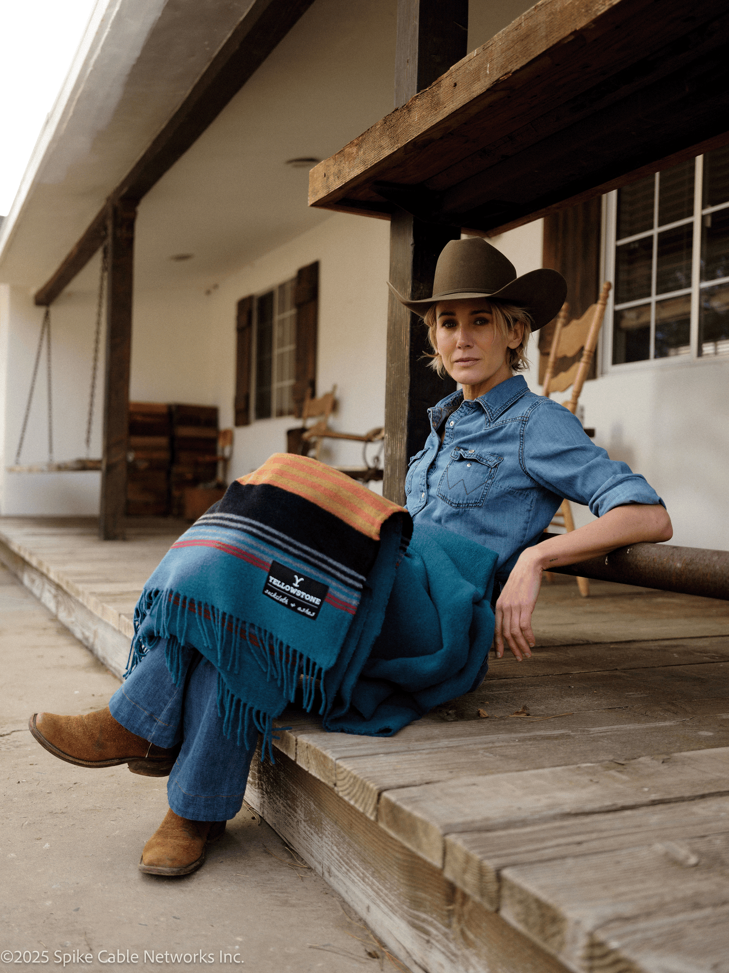 Woman in cowboy hat and blue denim outfit sitting on a wooden porch with a blanket.