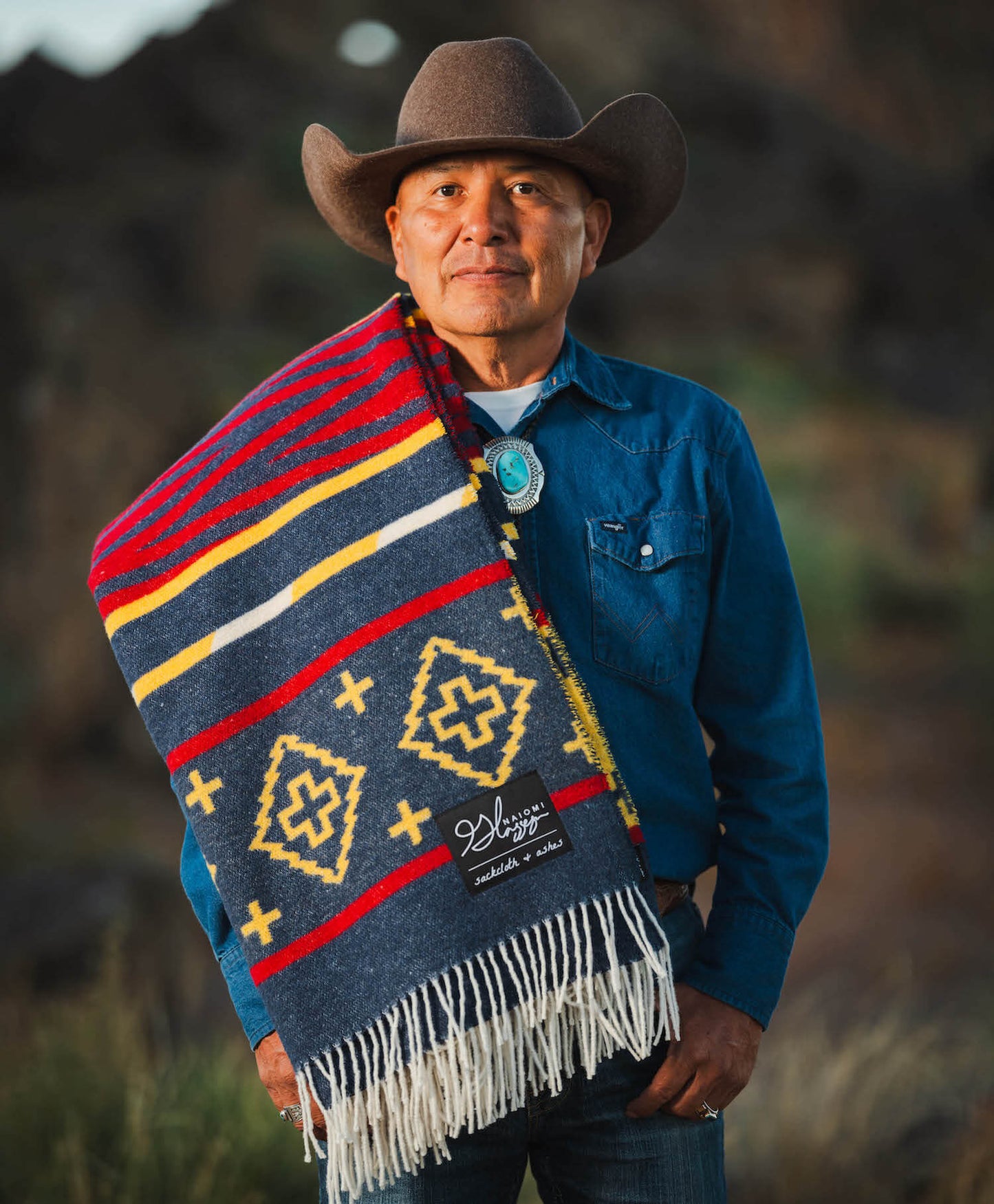 Man wearing a colorful patterned blanket and cowboy hat in a natural setting