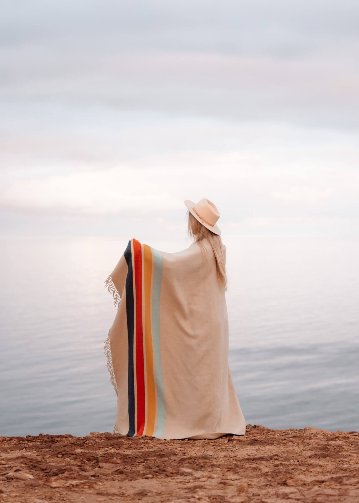 Person wearing a blanket with colorful stripes and a hat, standing on a rocky surface with a blurred background.