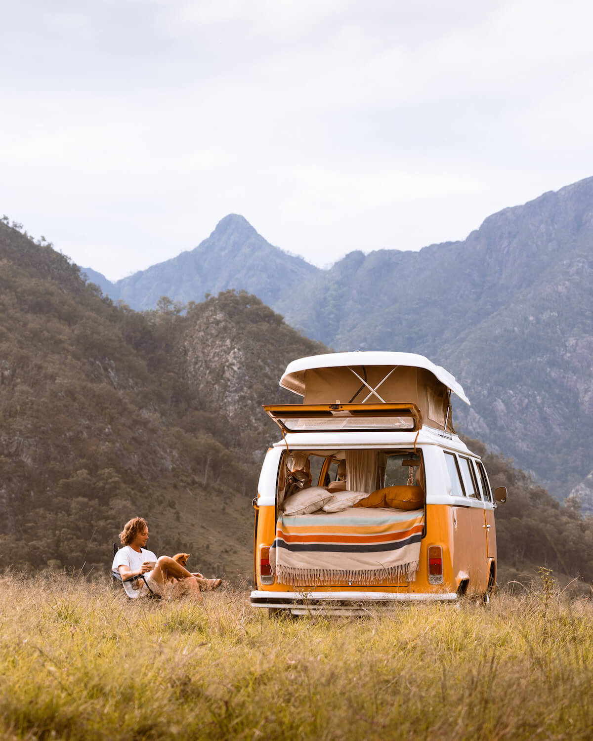 Person sitting in a field with a camper van featuring an open roof in a mountainous landscape