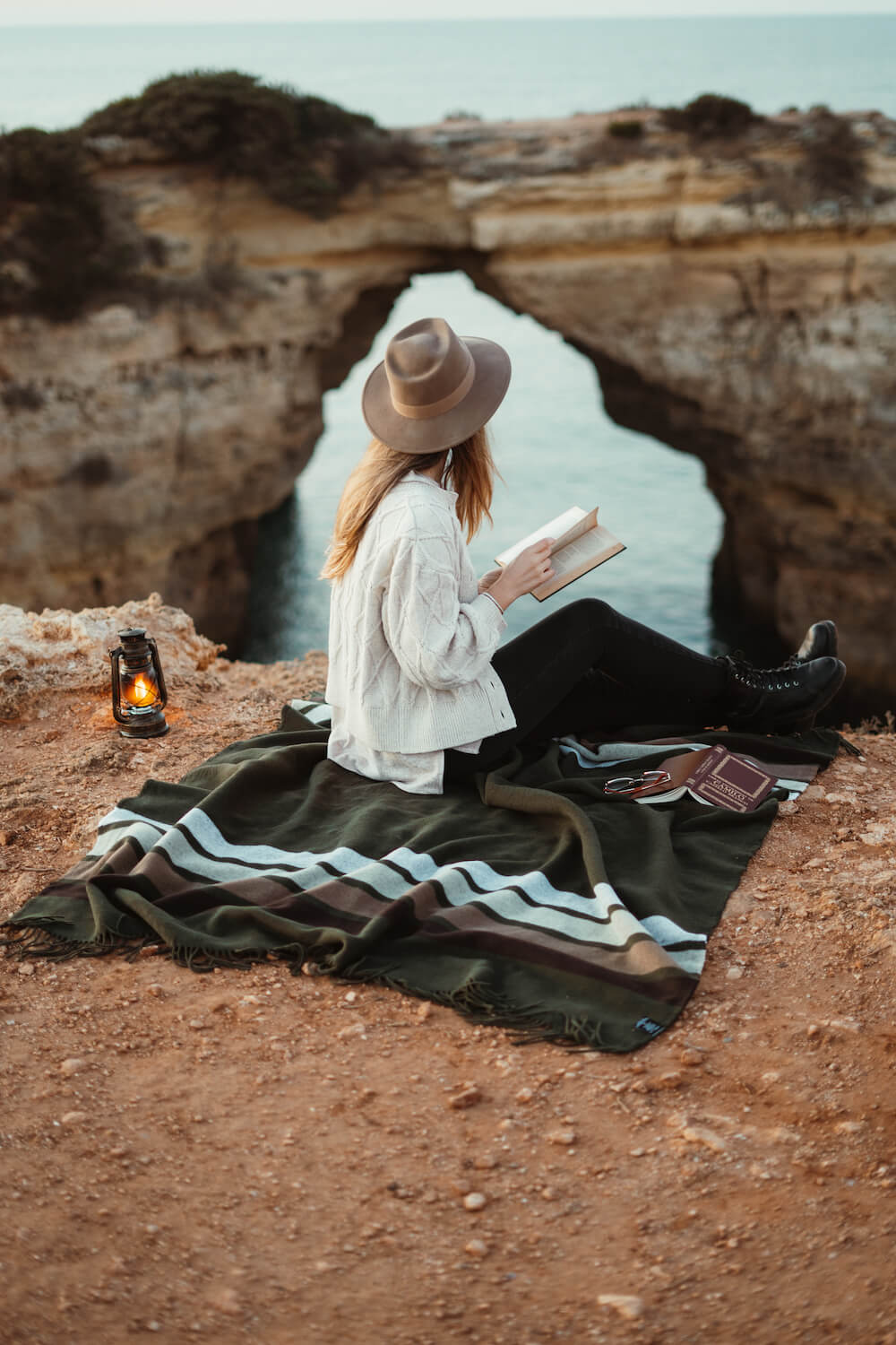 Woman reading a book on a blanket by a natural rock formation with a body of water.