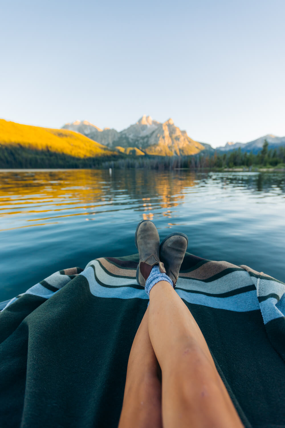 Person's feet with brown shoes and blue socks on a blanket by a lake with mountains in the background