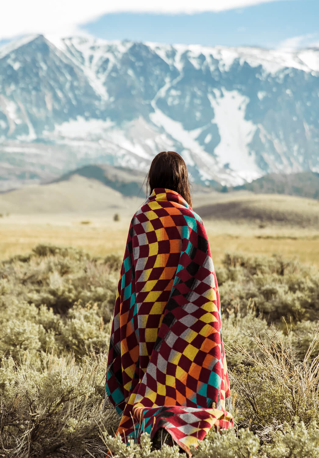 Person wrapped in a colorful blanket standing in a field with mountains in the background