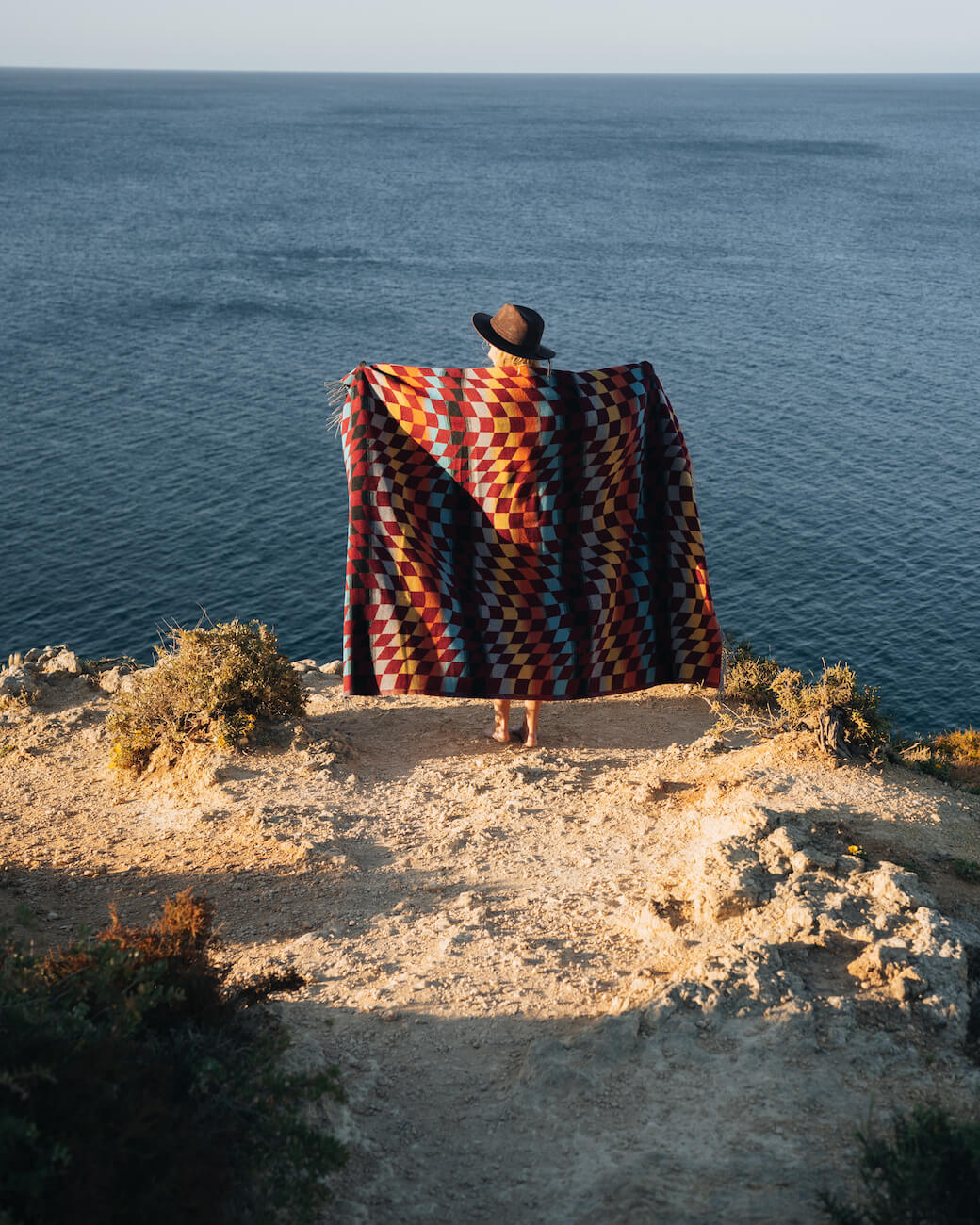 Person wrapped in a colorful blanket standing on a cliff overlooking the ocean.