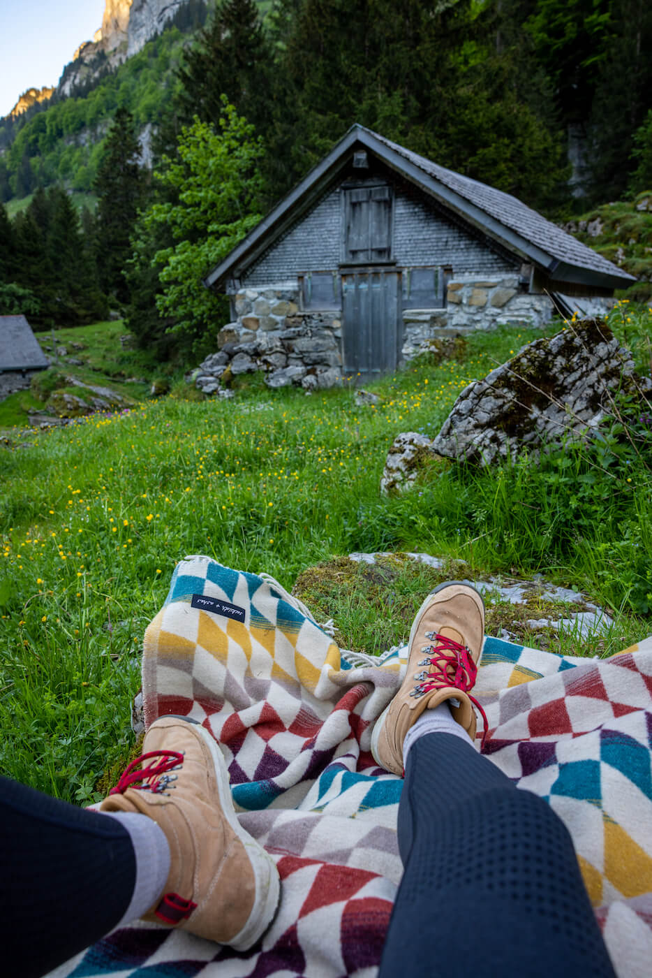 Person sitting on a colorful blanket with feet visible, wearing red shoes, in a scenic mountain setting with a stone cabin.