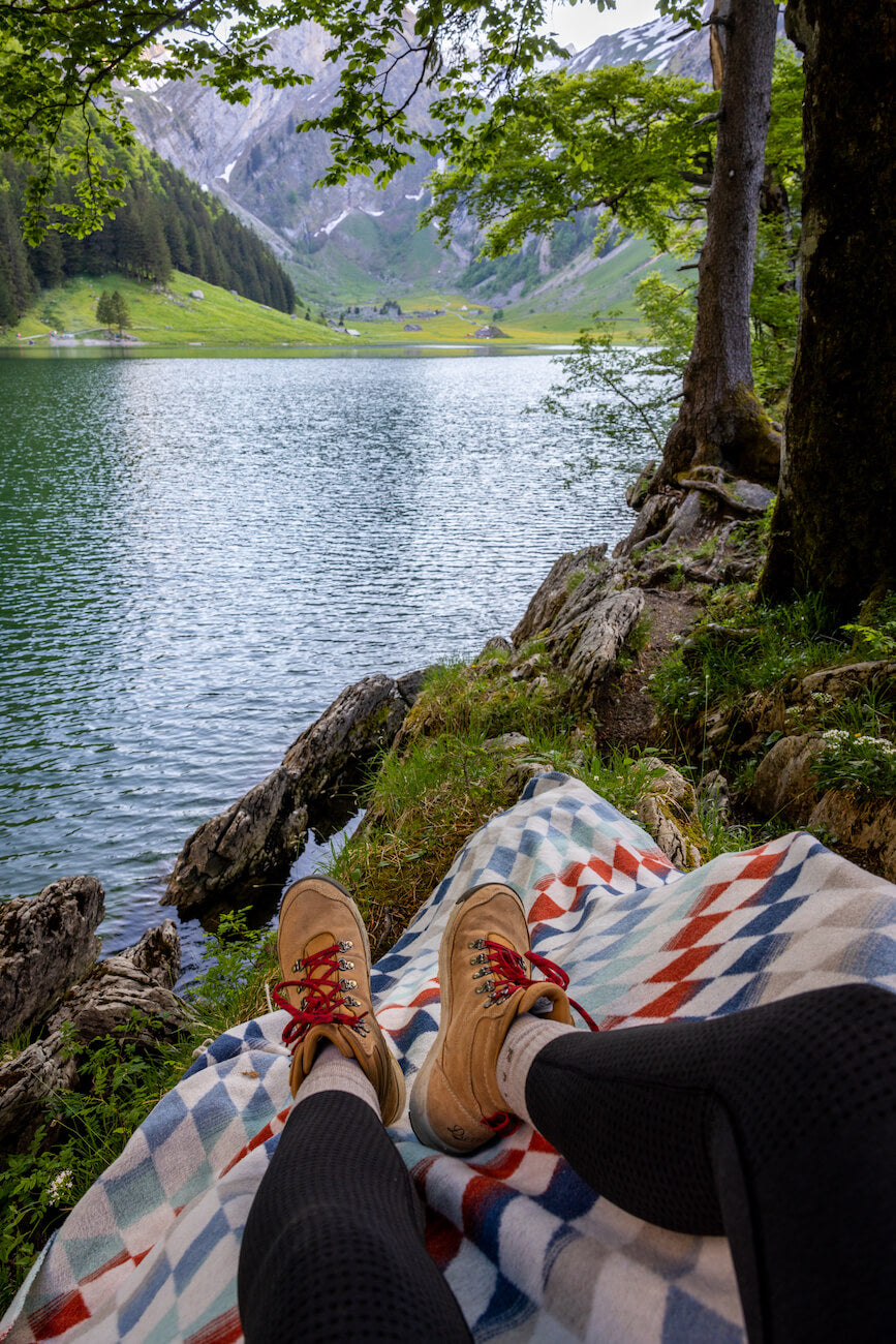 Person sitting on a colorful blanket by a lake with mountains in the background