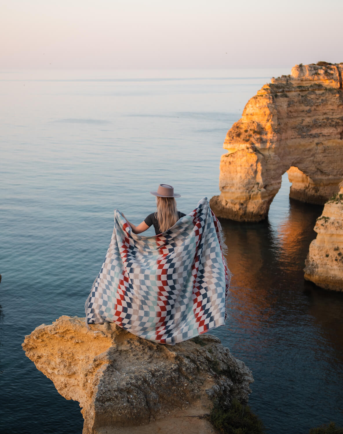 Person holding a patterned blanket on a rocky cliff overlooking the ocean.