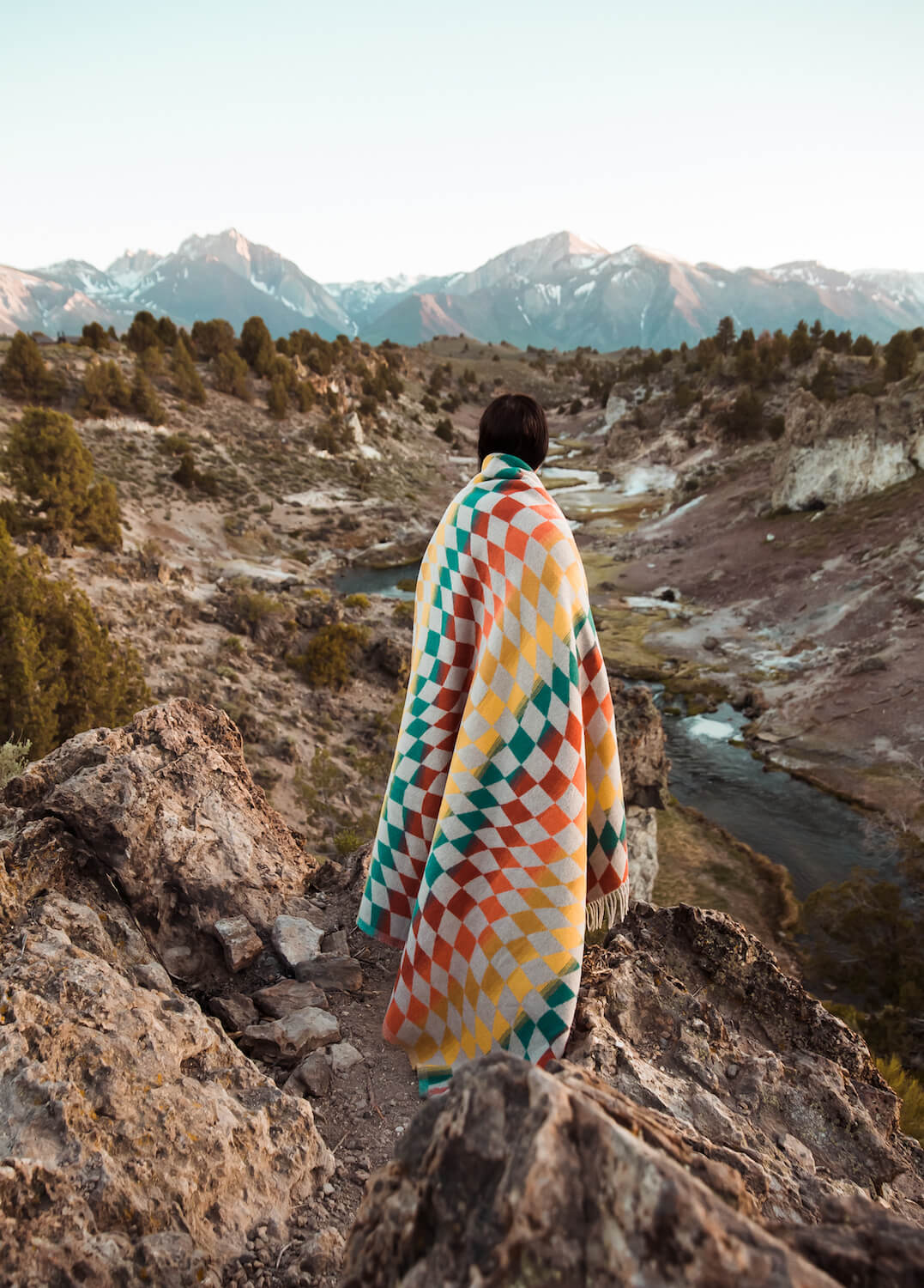 Person wrapped in a colorful diamond patterned blanket standing on a rocky mountain with a scenic view of mountains and a river.
