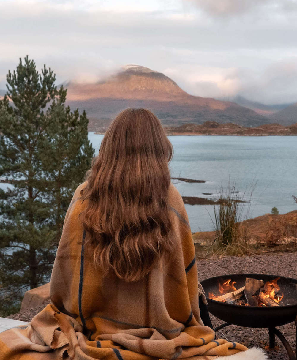 Person wrapped in a blanket sitting by a fire pit with a scenic view of a lake and mountain.