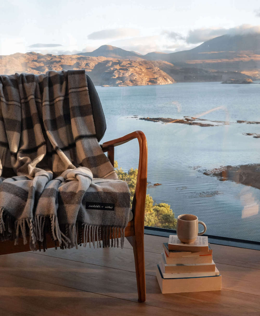 Chair with a plaid blanket and a cup of coffee on books, overlooking a lake and mountains.