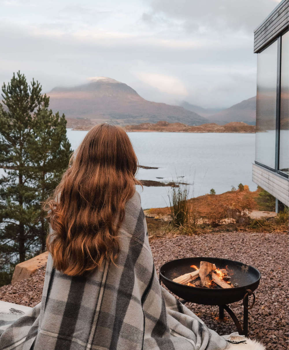 Person wrapped in a blanket by a fire pit with a scenic view of a lake and mountains.
