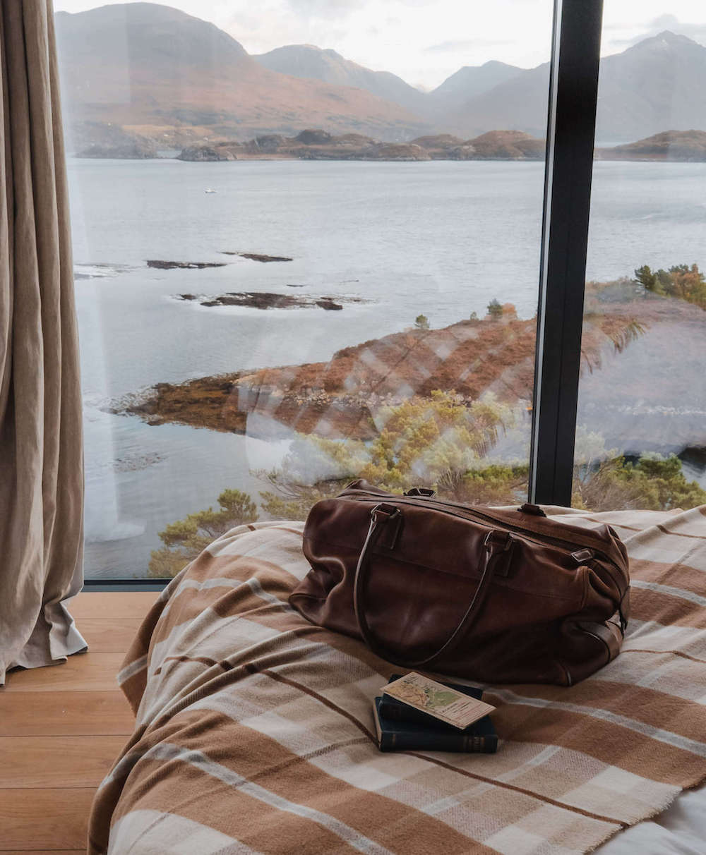 Brown leather bag on a bed with a scenic view of a lake and mountains through large windows.