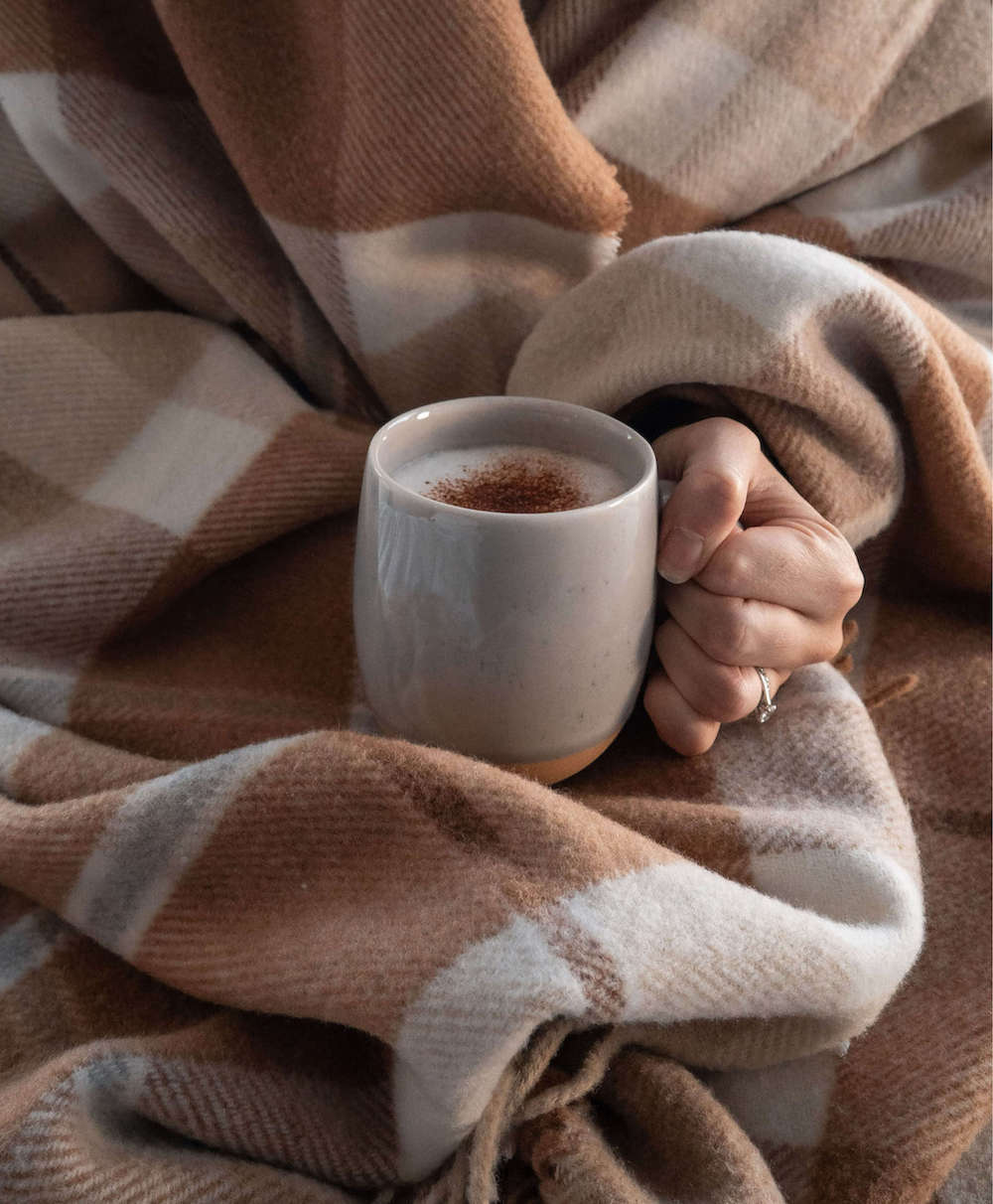 Hand holding a mug of hot chocolate wrapped in a cozy plaid blanket