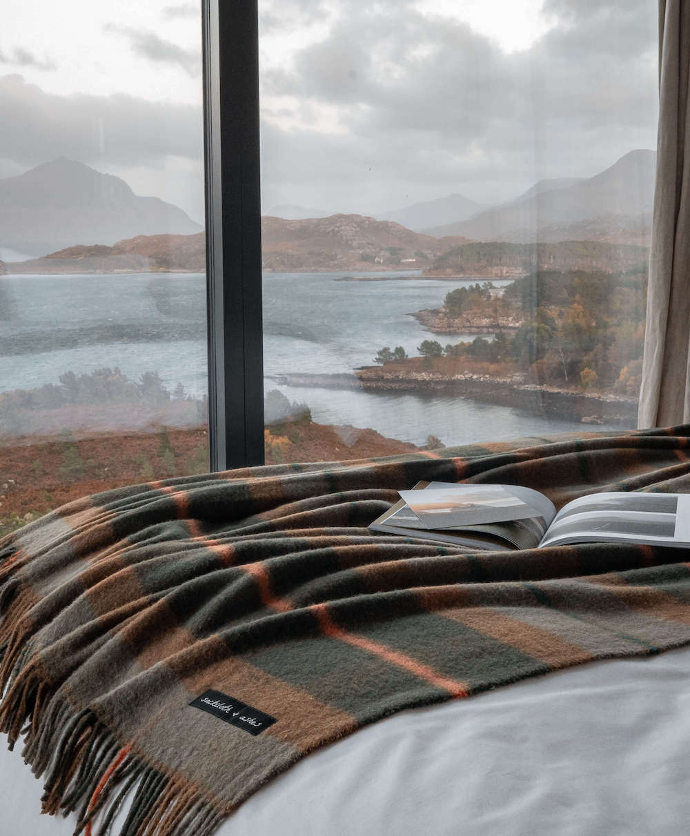 Plaid blanket draped over a bed with a scenic view of a lake and mountains through large windows.