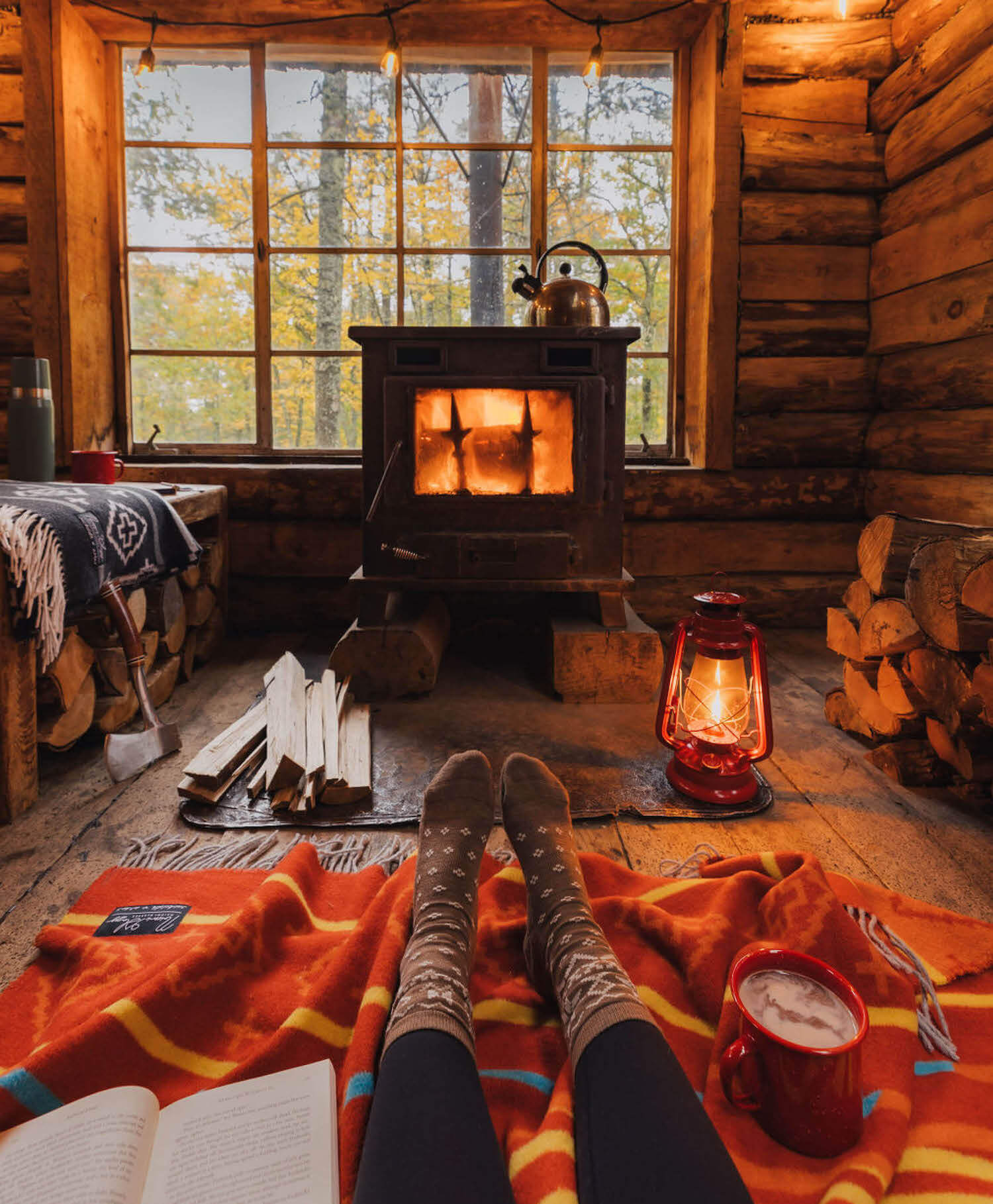Log cabin interior with a lit stove, lantern, and person's feet on an orange blanket.