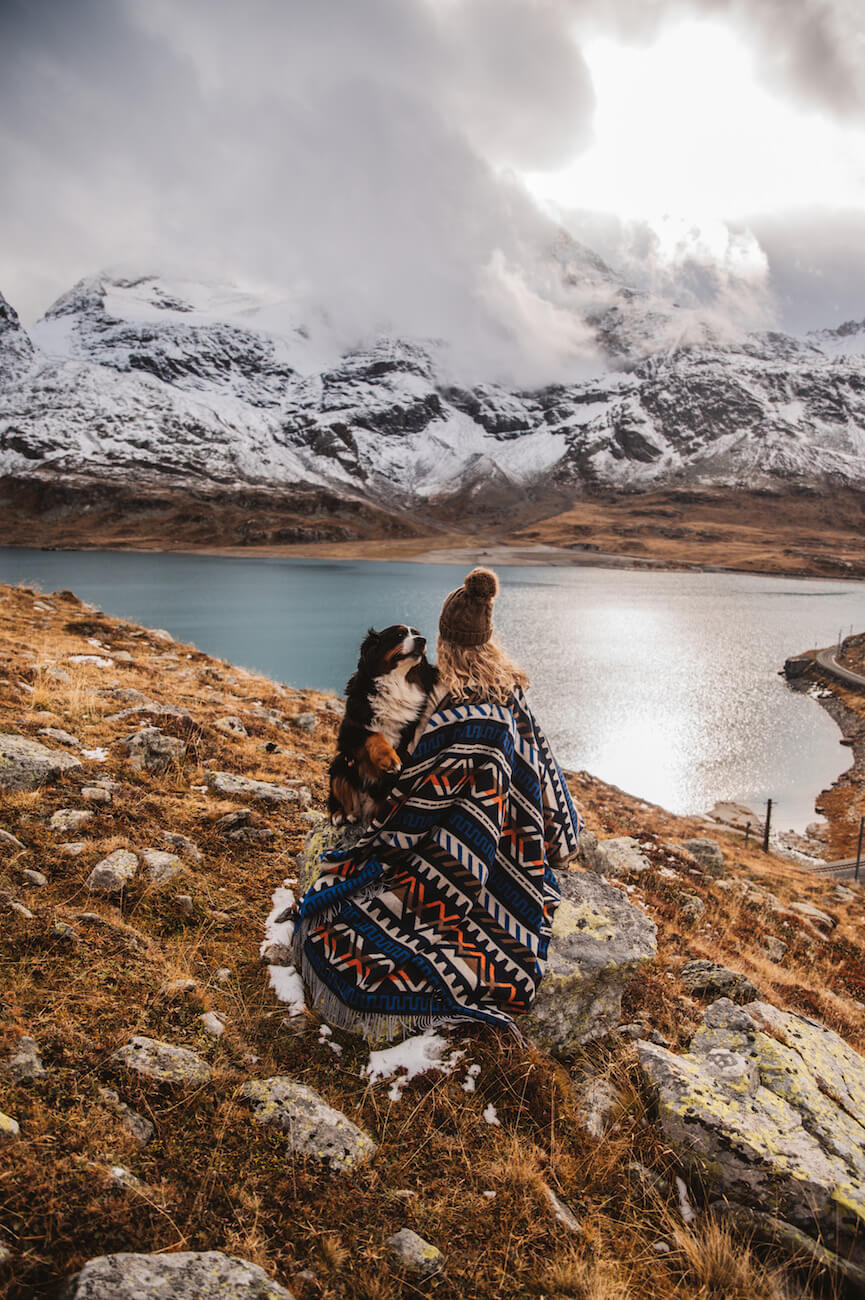 Person and dog sitting on a blanket overlooking a mountain lake with snow-capped peaks.