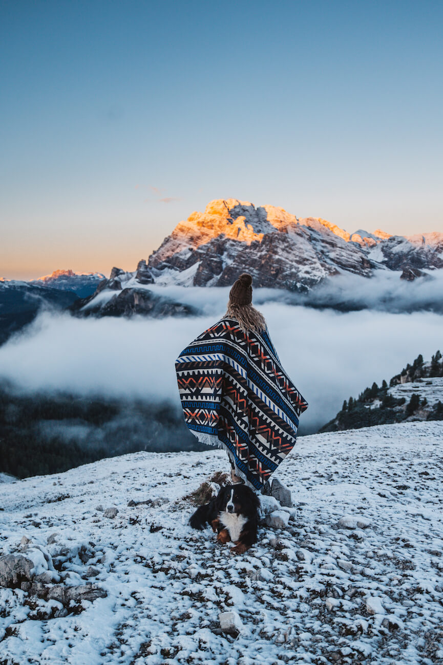 Person wrapped in a patterned blanket with a dog on a mountain top at sunset.