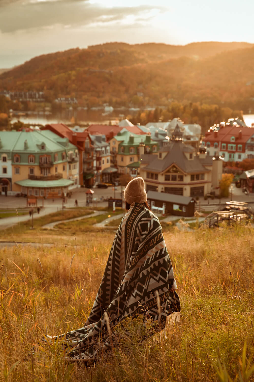 Person wrapped in a blanket sitting in a field with a town and mountains in the background