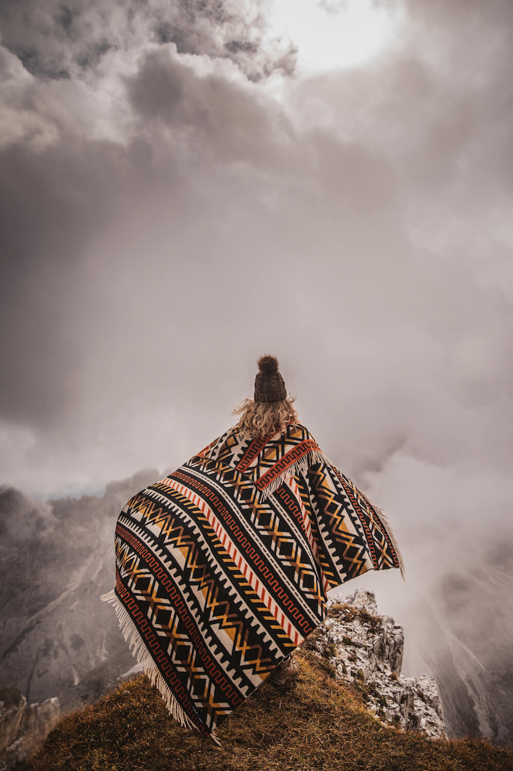 Person wearing a patterned poncho standing on a mountain with clouds in the background
