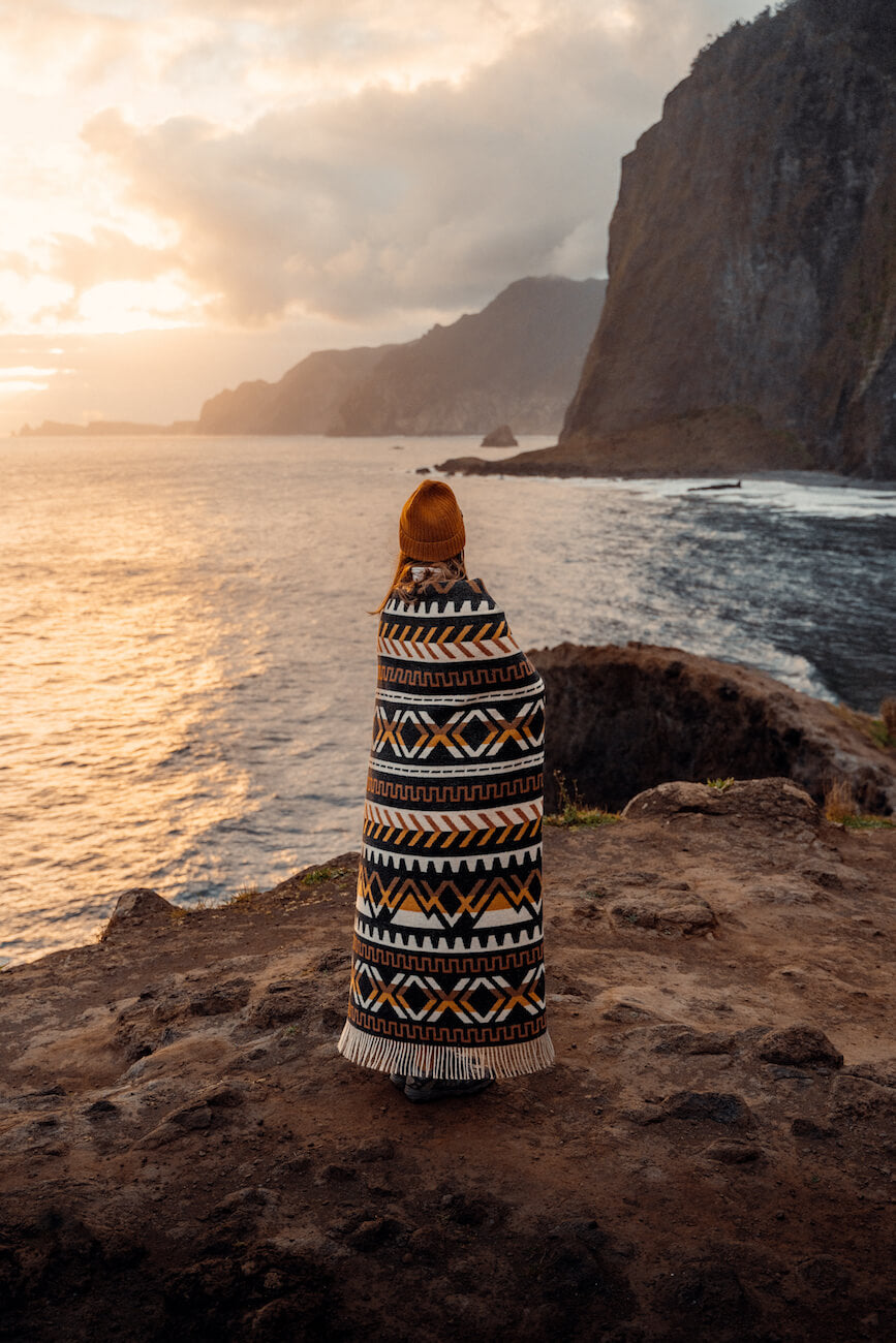 Person wrapped in a patterned blanket standing on a rocky cliff overlooking the ocean at sunset.