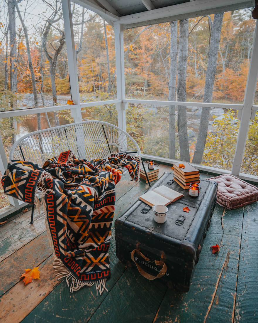 Screened porch with autumn decor, including a patterned blanket, books, and candles, with trees and water in the background.