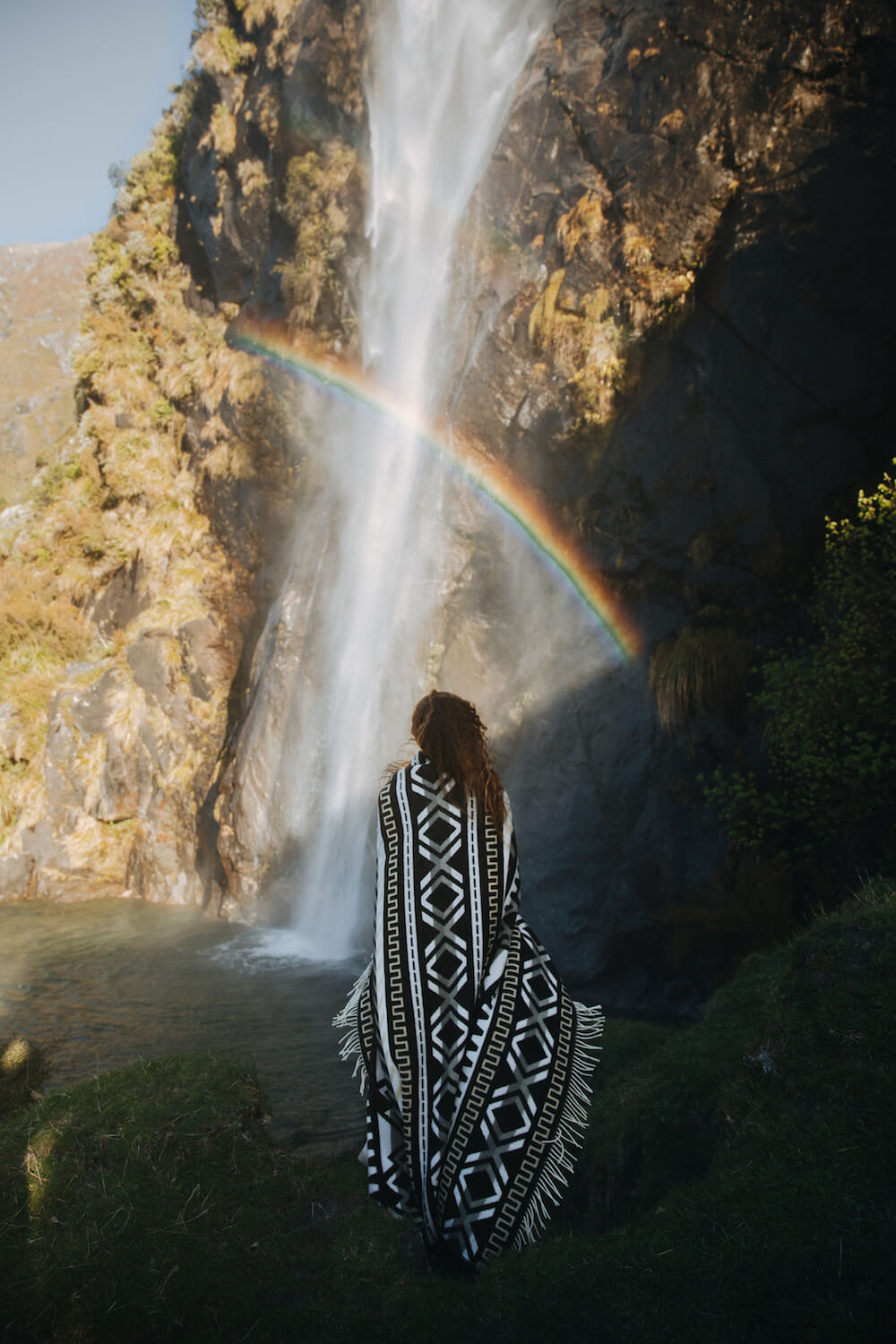 Person in a patterned poncho standing in front of a waterfall with a rainbow, surrounded by rocky cliffs and greenery.