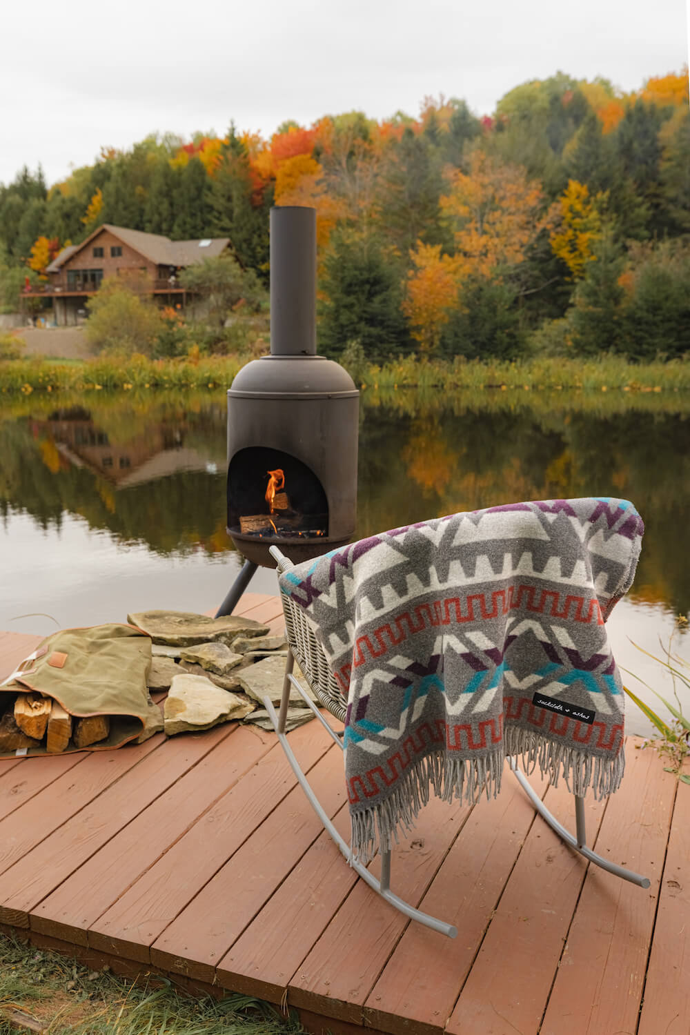 Outdoor fireplace on a wooden deck with a colorful blanket and firewood, surrounded by autumn trees.