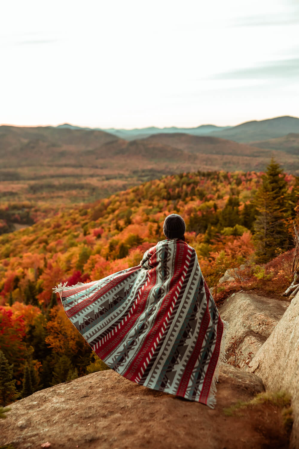Person standing on a rock overlooking a scenic view of mountains and trees with autumn colors.