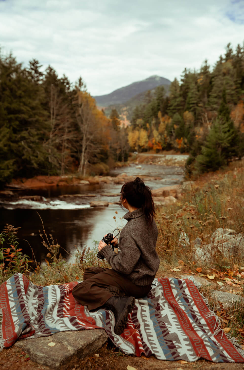 Person sitting on a colorful blanket by a river with trees and mountains in the background
