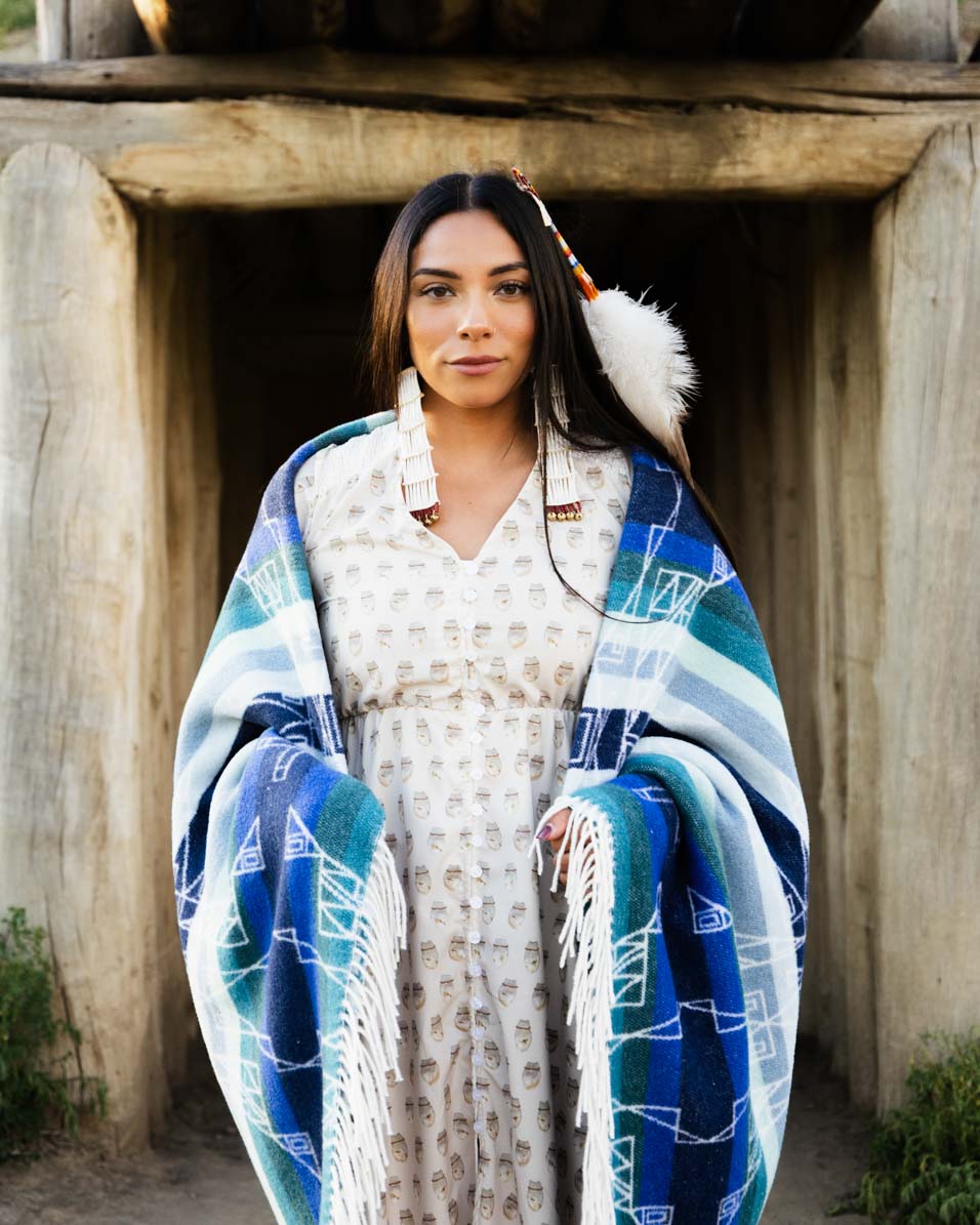 Woman wrapped in a blue and green patterned blanket standing in front of wooden structures.