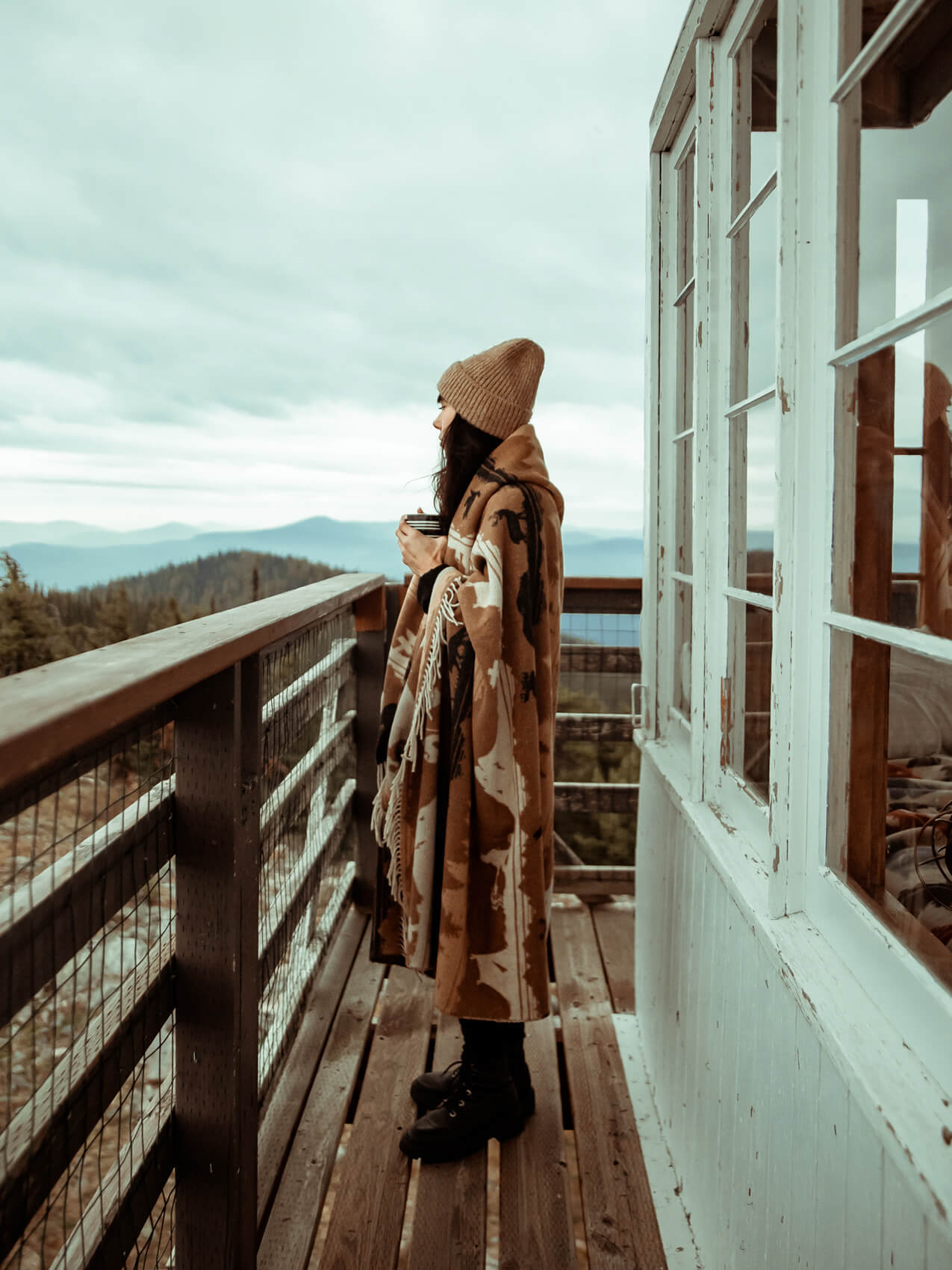 Person standing on a wooden balcony overlooking a scenic view with mountains and trees.