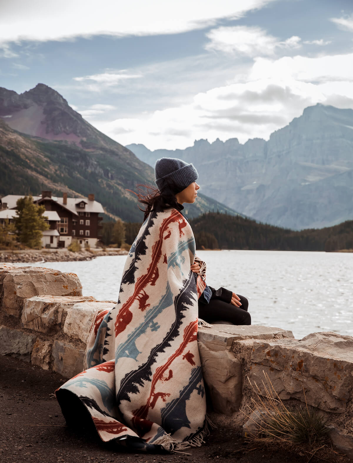 Person wrapped in a patterned blanket sitting by a lake with mountains in the background